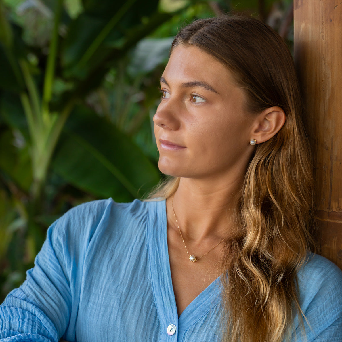 Woman in a blue shirt leaning against a wooden wall with greenery in the background