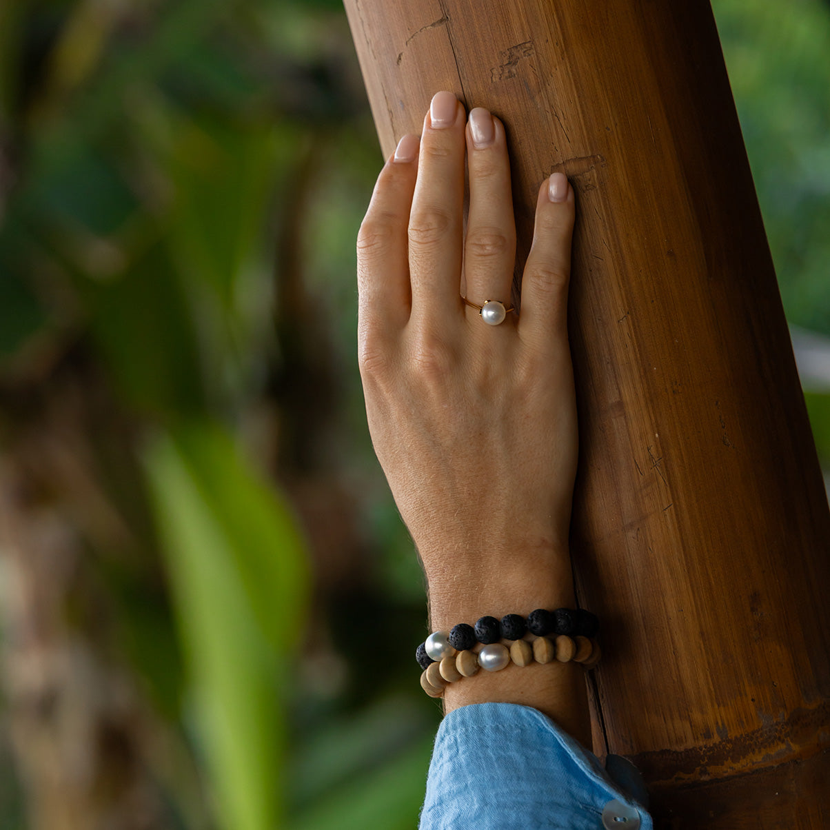 Hand with a pearl ring and beaded bracelet holding a wooden surface, with a blurred natural background