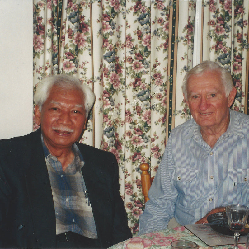Two ederly men sitting at a dinning table with flora curtins in the back.