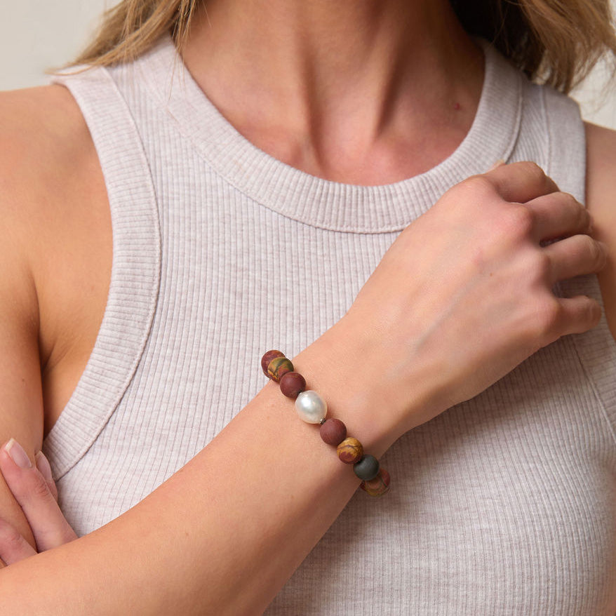 Person wearing a bracelet with red and orange beads and a pearl on a neutral background