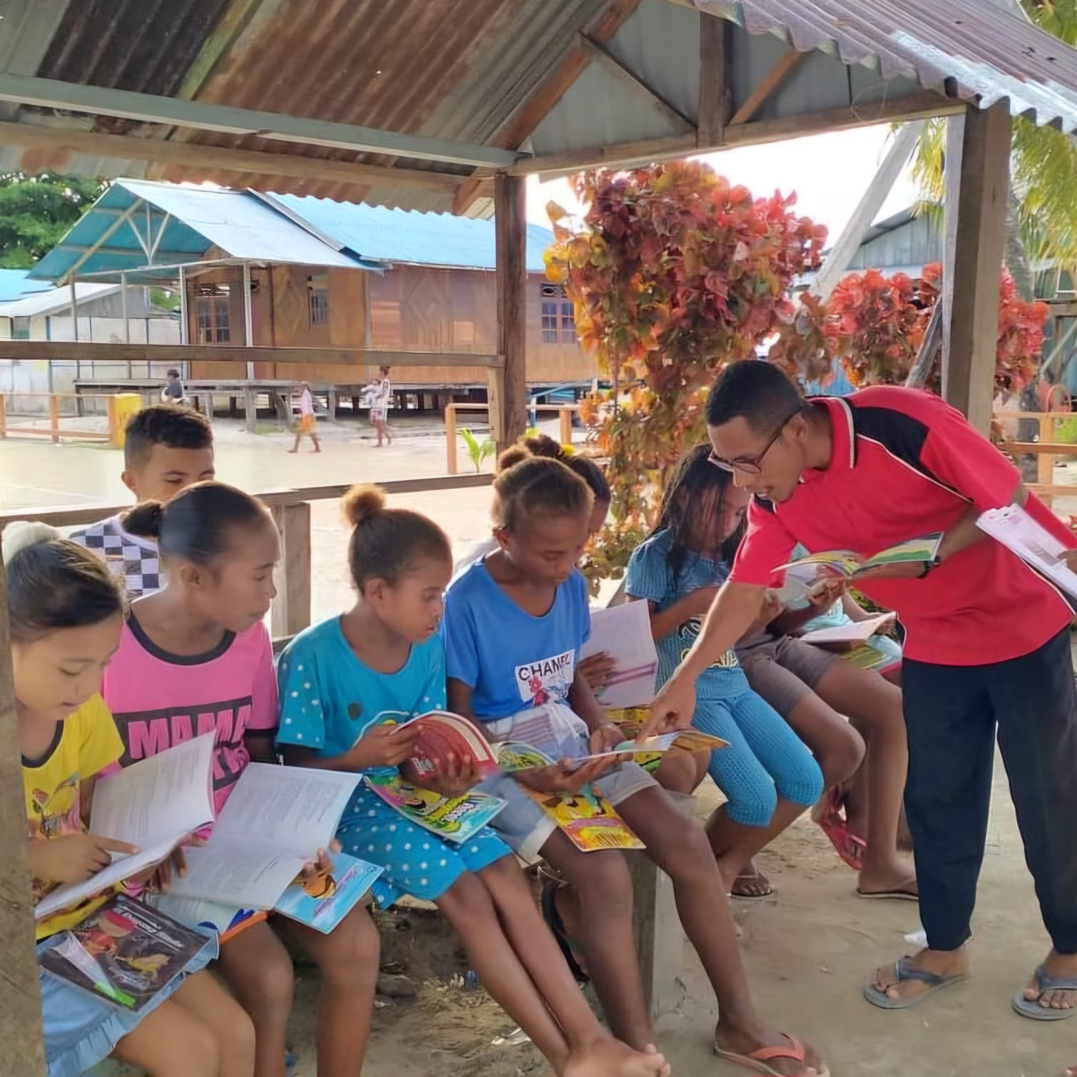 Children sitting under a wooden shelter, reading books with an adult nearby.